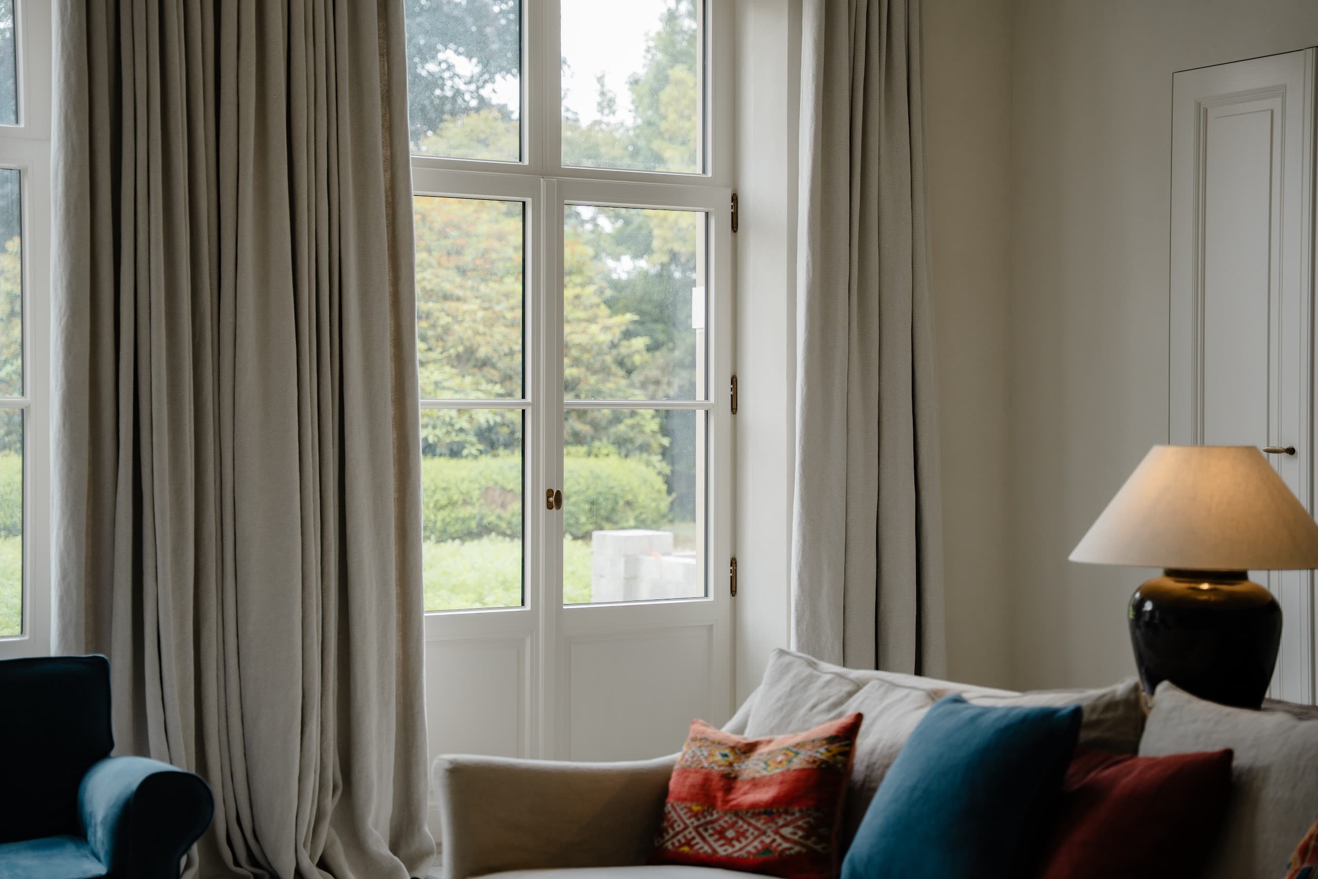 Woonkamer met loungebank, schemerlamp en overgordijnen langs een hoog raam